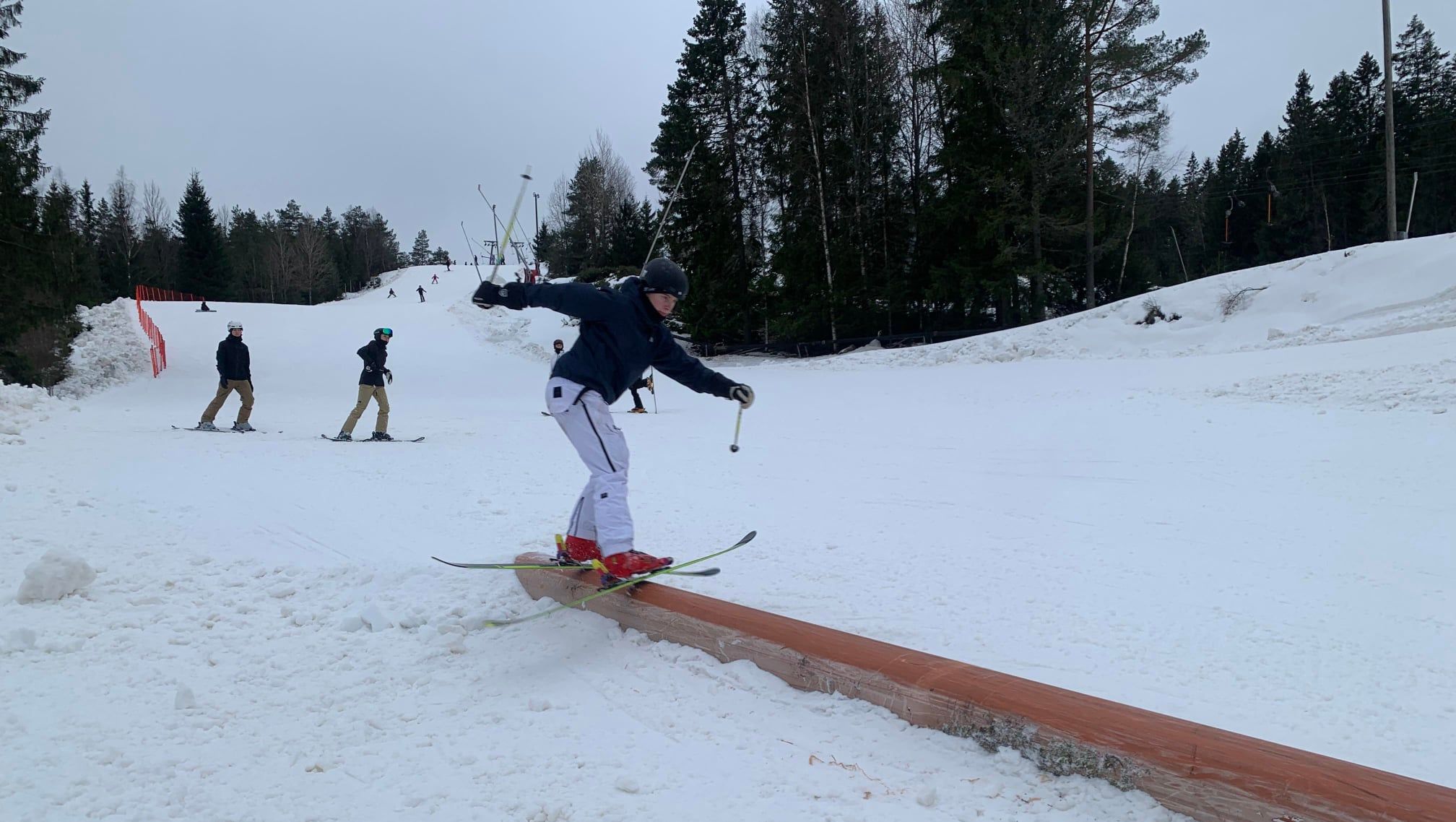 Skiløper som balanserer på en rail i terrengparken, med andre kjørere i bakken bak.